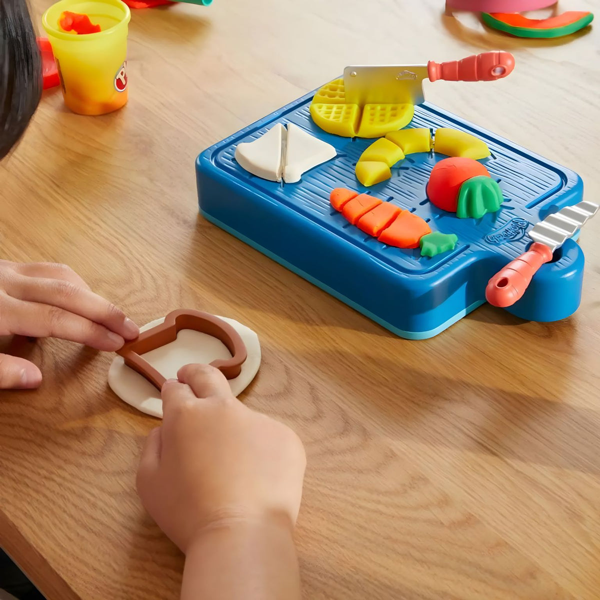 Criança a usar um cortador de pão junto da tábua com alimentos de massa e facas de brincar