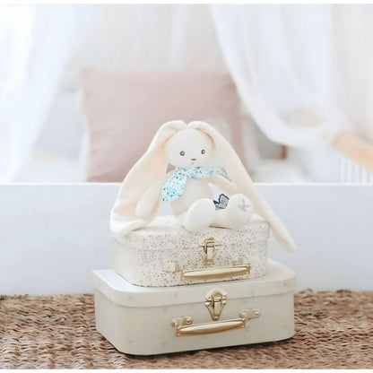 Peluche Coelho Creme 25 cm da Kaloo sentado sobre malas num quarto